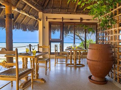 Área de restaurante abierta con mesas y sillas de madera, vista al mar y palmeras.