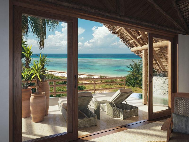 Blick durch große Glastüren auf Terrasse mit Liegestühlen, Palmen und blauem Meer im Hintergrund.