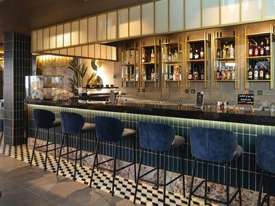 Bar d'hôtel élégante avec comptoir sombre, tabourets bleus et étagères lumineuses.