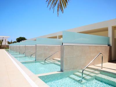 Zone de piscine moderne avec cloisons en verre et escaliers, entourée de bâtiment blanc et ciel bleu.
