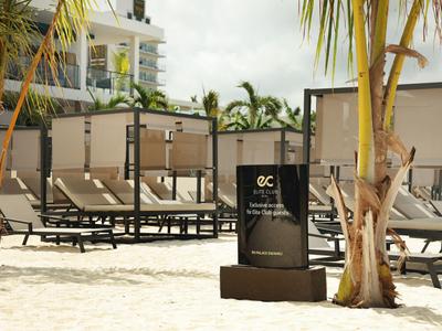 Plage de sable avec chaises longues et panneau devant des palmiers près d'une piscine d'hôtel