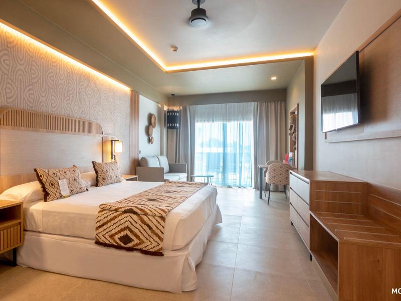 Nowoczesny pokój hotelowy z łóżkiem podwójnym, telewizorem z płaskim ekranem, częścią wypoczynkową i balkonem z widokiem na morze.