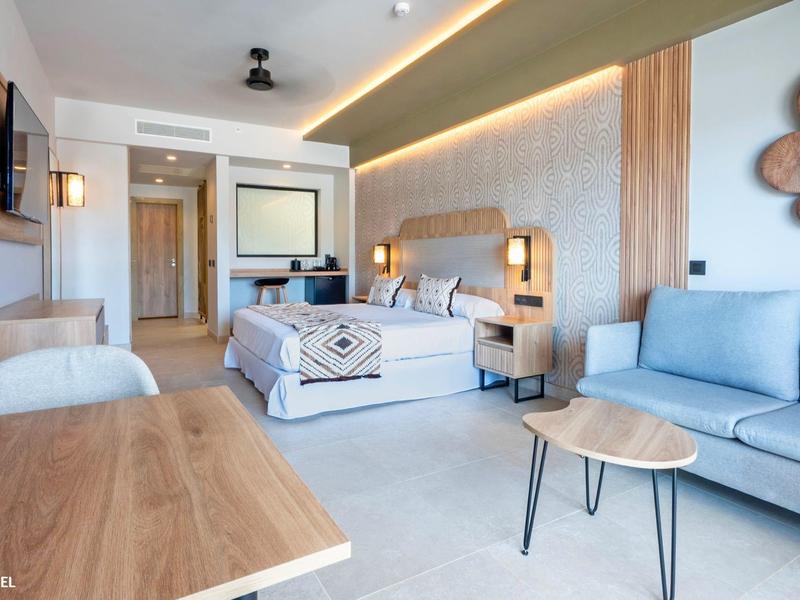 Nowoczesny pokój hotelowy z łóżkiem, sofą, stołem i dekoracją ścienną w jasnych kolorach.