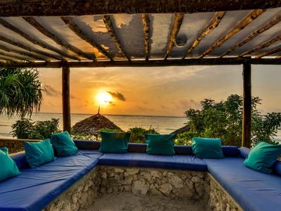 Comfortabele zithoek met blauwe kussens onder een houten dak bij zonsondergang aan zee.