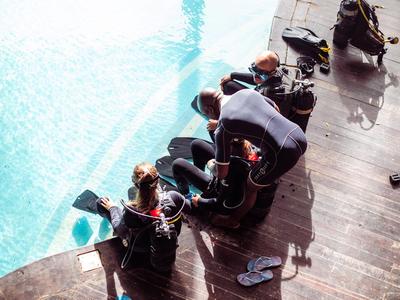 Drie mensen zitten aan de rand van het zwembad met duikpakken en uitrusting naast hen.