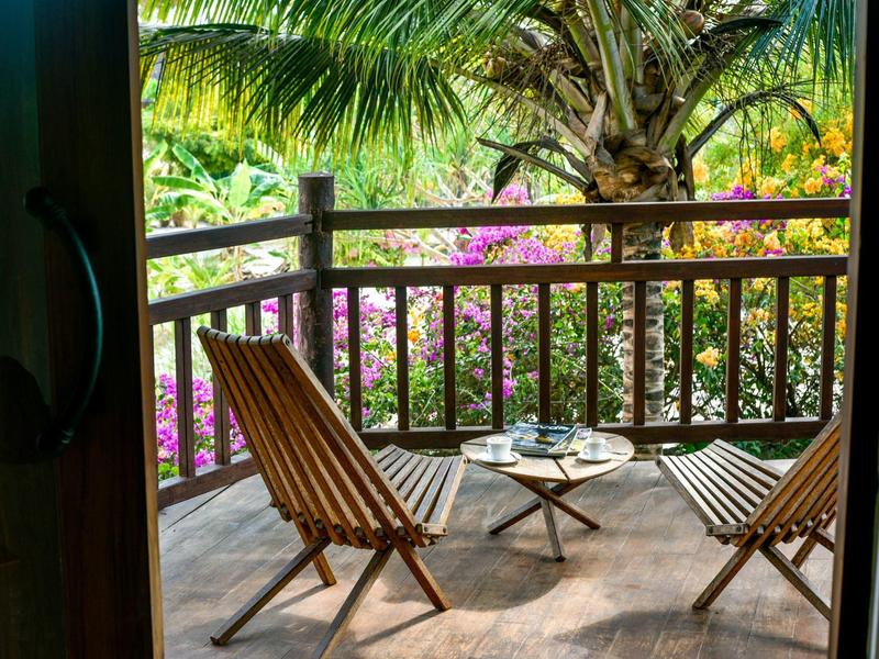Zwei Holzstühle und ein kleiner Tisch auf einer Veranda mit Blick auf tropische Pflanzen.
