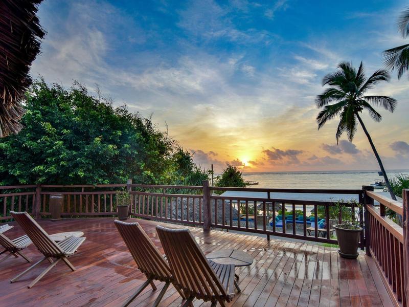 Holzterrasse mit Liegestühlen, Blick auf das Meer bei Sonnenuntergang, umgeben von Palmen.