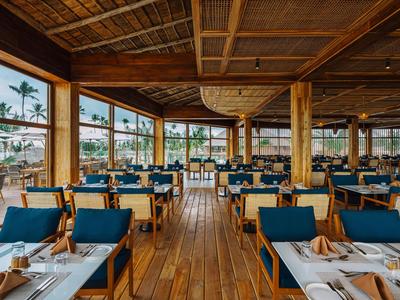 Grande ristorante con mobili in legno e sedie blu, lati aperti con vista sulle palme.