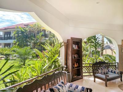 Zastřešená terasa s dřevěným nábytkem s výhledem na tropickou zahradu a hotelové budovy.