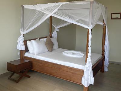 Schlafzimmer mit Himmelbett aus Holz, weißem Moskitonetz und schlichtem Nachttisch.