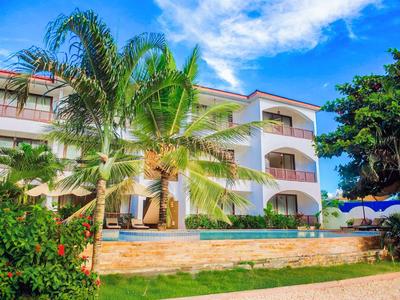 Třípodlažní hotelová budova s palmami a bazénem pod modrou oblohou.