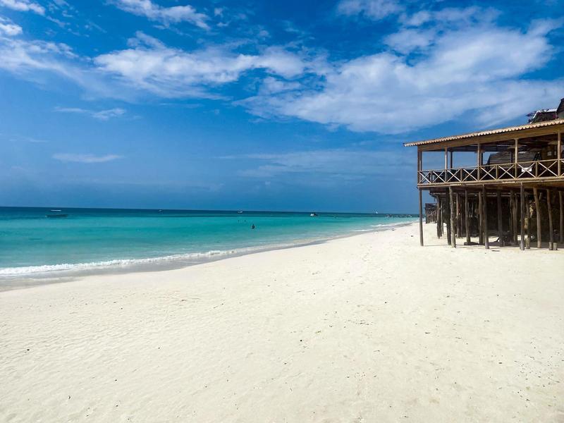 Ein weißer Sandstrand mit klarem türkisfarbenem Wasser und einem über Stelzen gebauten Holzhaus.