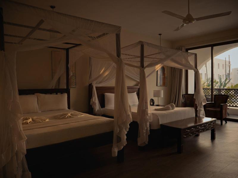 Elegantes Hotelzimmer mit Baldachinbetten, Holzmöbeln und Blick auf eine Terrasse bei gedämpftem Licht.