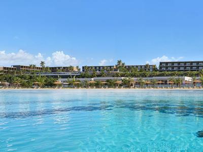 Breites Hotel mit moderner Architektur am Strand und klarem, blauem Wasser im Vordergrund.