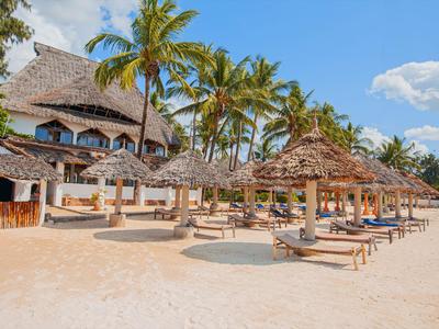 Beach hotel with thatched roofs, sun loungers, and palm trees under a blue sky.