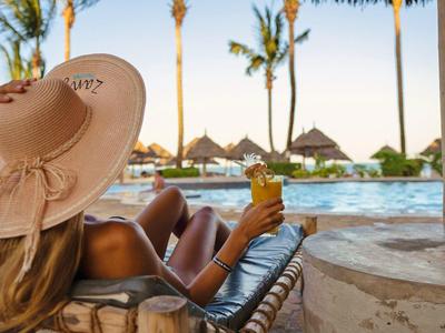 Mujer con sombrero de paja relajándose en la piscina con bebida tropical y palmeras de fondo.