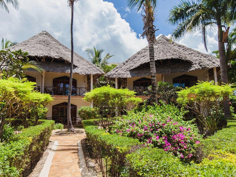 Resort tropical con bungalows de techo de paja, vegetación exuberante y palmeras bajo cielo azul.