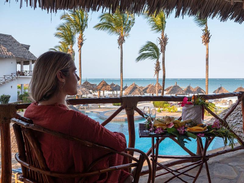 Mujer relajándose con vista a la piscina, palmeras y playa al atardecer en un hotel tropical.