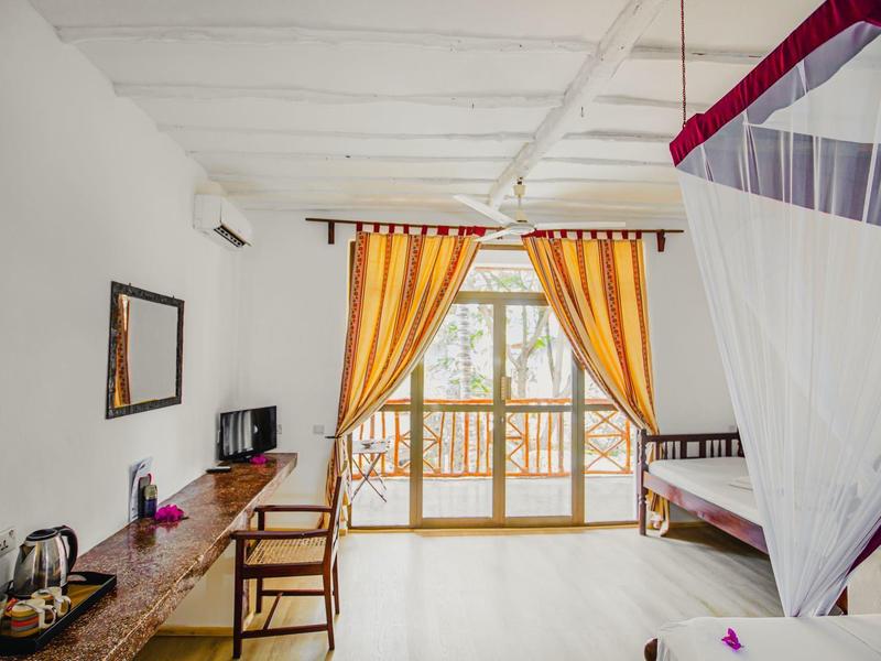 Habitación de hotel luminosa con balcón, cortinas, cama con mosquitera, escritorio y silla.