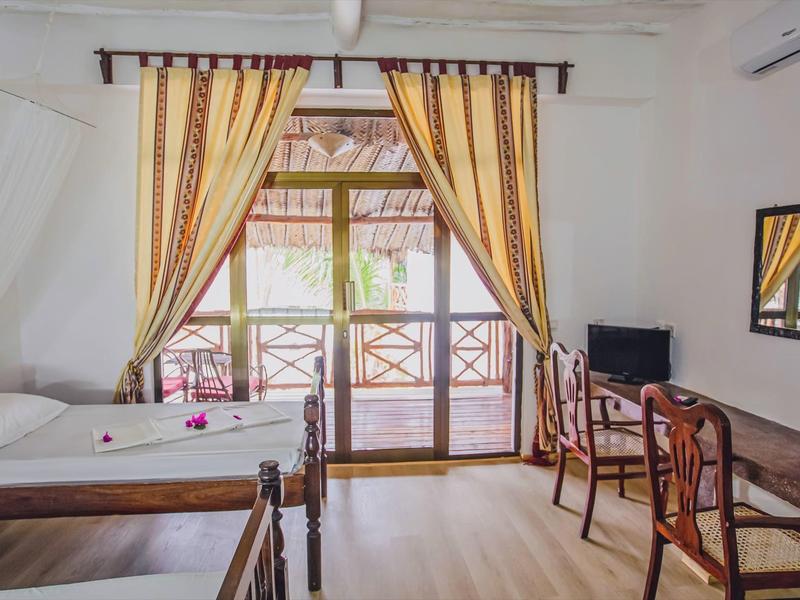 Habitación de hotel luminosa con dos camas, escritorio, sillas y balcón con barandilla de madera.