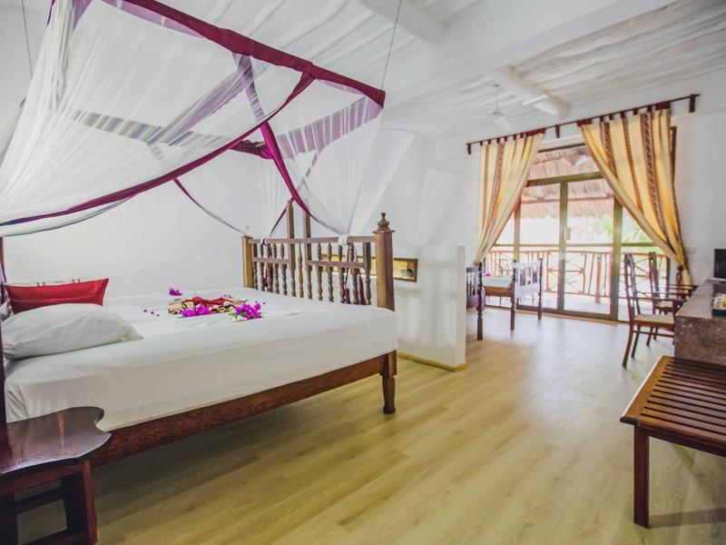 Habitación luminosa con muebles de madera, cama doble con mosquitero y acceso a balcón con cortinas.