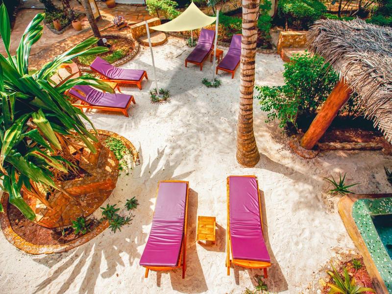 Sillas coloridas sobre arena blanca, rodeadas de palmeras y una pequeña piscina en un hotel tropical.