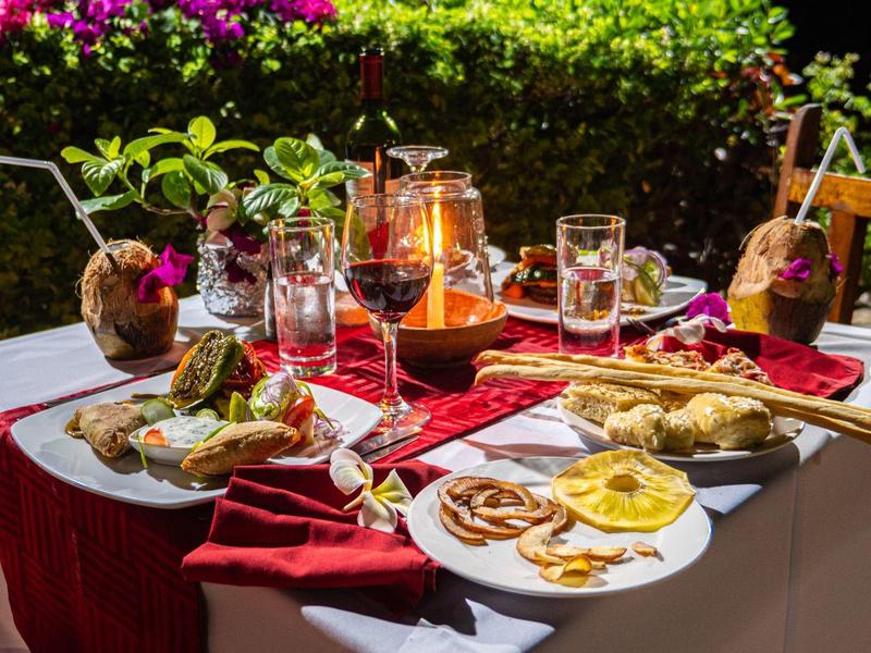 Mesa al aire libre con bebidas y platos variados sobre mantel blanco y rojo, rodeada de vegetación.