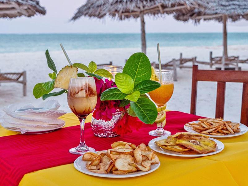 Platos con pasteles y dos bebidas sobre una mesa con manteles coloridos en la playa.