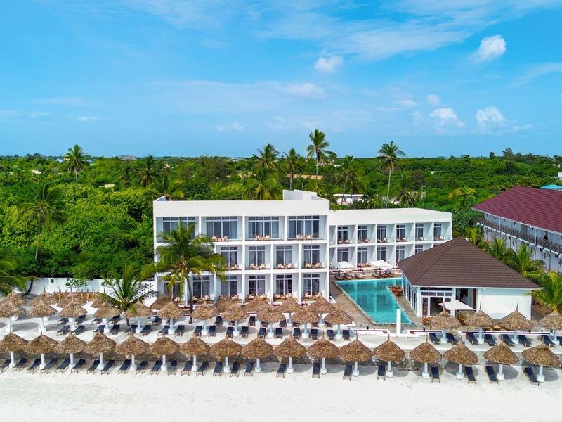 Strandresort mit weißem Gebäude, Pool und vielen Sonnenschirmen am Sandstrand unter blauem Himmel.