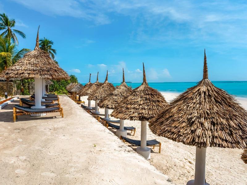 Reihe von Strohdach-Sonnenschirmen und Liegestühlen am sandigen Strand bei klarem Himmel.