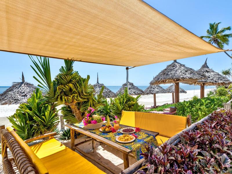 Bunte Sitzgruppe mit Essen unter Sonnensegeln am tropischen Strand mit Palmen.