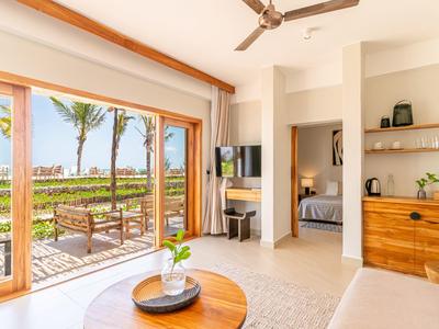 Helles Hotelzimmer mit Terrasse, Holzmöbeln und Blick auf Palmen und Garten.