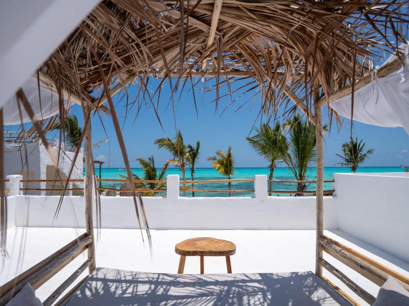 Blick von einer überdachten Terrasse mit Strohdach auf Palmen und das blaue Meer.