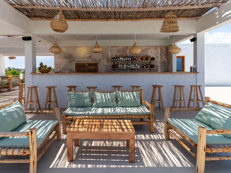 Outdoor-Lounge mit Holzmöbeln, Kissen und Bar unter einem schattigen Dach.