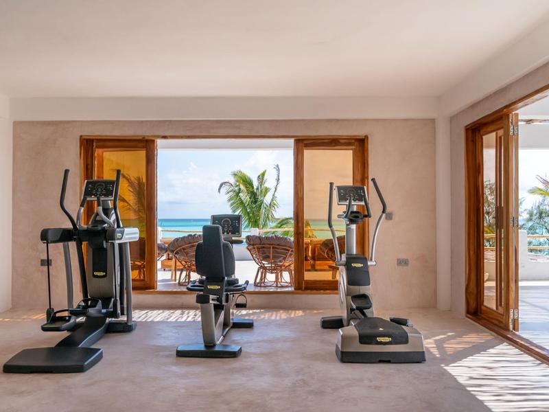 Fitnessraum mit drei Trainingsgeräten vor großen Fenstern mit Meerblick.