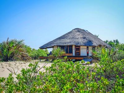 Bungalow mit Strohdach am Strand, umgeben von grünem Gebüsch und blauem Himmel.