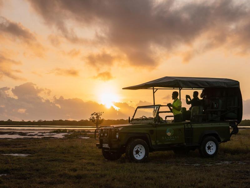 Jeep da safari al tramonto in un paesaggio erboso con acqua sullo sfondo.