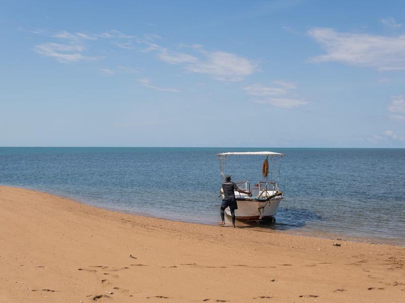 Una persona accanto a una piccola barca al margine di una spiaggia calma sotto un cielo azzurro.