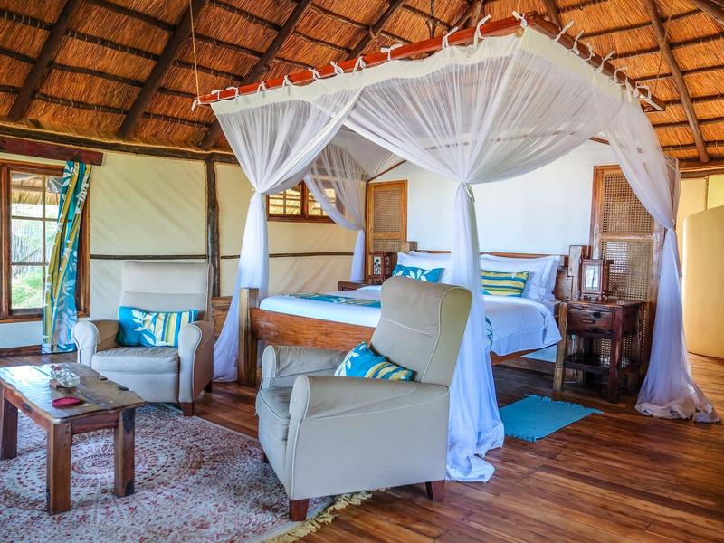 Camera d'hotel accogliente con letto a baldacchino, poltrona e pavimento in legno sotto un tetto di paglia.