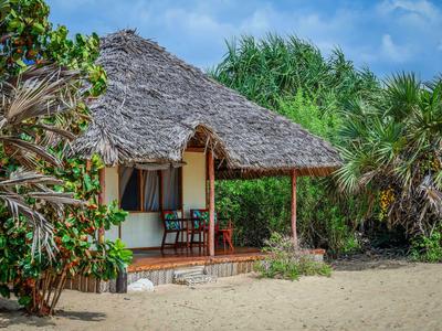 Kleines Hotelzimmer mit Strohdach und Terrasse am Sandstrand, umgeben von Palmen.
