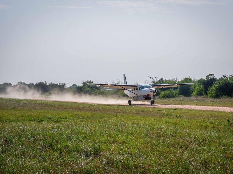 Pequeño avión de hélice aterriza en una pista de césped con nubes de polvo al fondo.